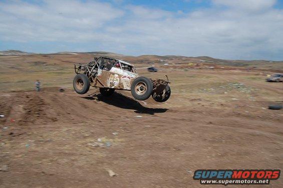 Rusty off-road buggy mid-air, white body with red accents, exposed roll cage, dirt track