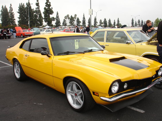 Yellow coupe with black hood stripes and aftermarket wheels parked at an outdoor car show. Another yellow car is parked next to it
