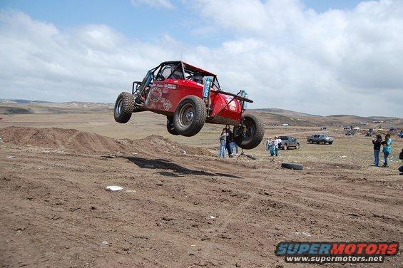 red off-road buggy mid-air over dirt track, people watching in background