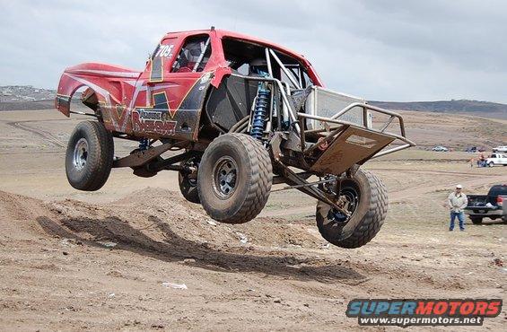Red off-road truck mid-air, custom roll cage, large tires, dirt track