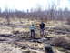 1996 Ford Bronco - Two individuals stand in a muddy, off-road terrain with sparse trees and dry brush surrounding them.