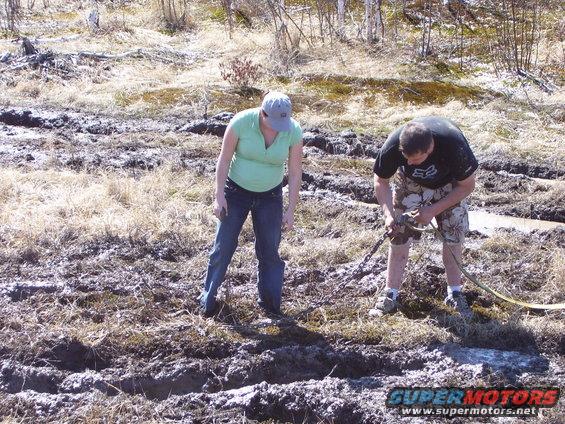 Two people are in a muddy, grassy field. One person is attaching a tow strap to something off-camera