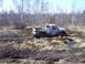 1996 Ford Bronco - A white pickup truck is stuck in a muddy, wooded area, with its tires sunk deep into the mire.