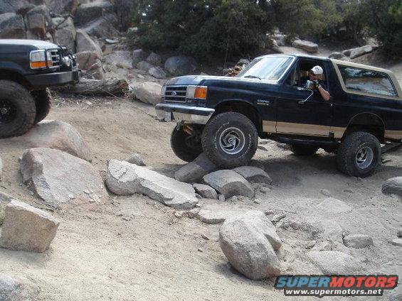 Two Ford Broncos off-roading on a rocky trail. The front Bronco is black with a tan top, and the second Bronco is partially visible on the left