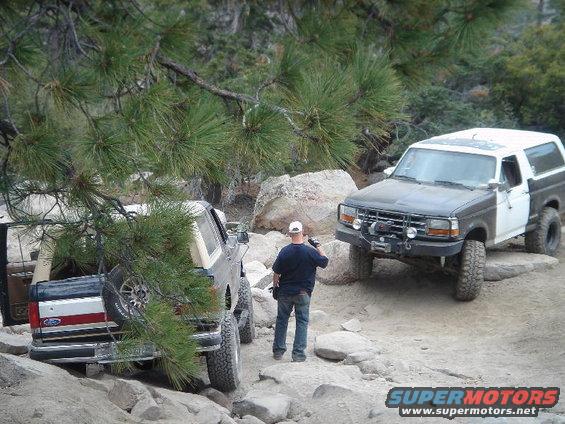 Two SUVs are navigating a rocky trail. One is a dark and white Bronco, the other is a dark Bronco with a spare tire on the back