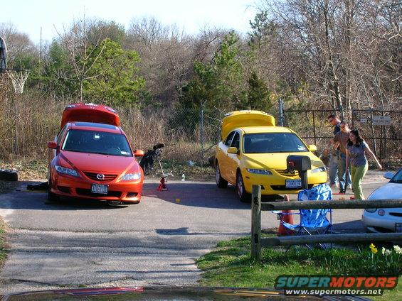 Two sedans, one red and one yellow, parked with hoods open. People stand near the yellow car