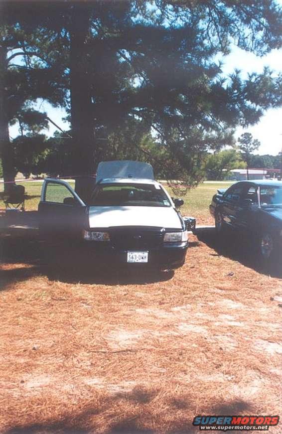 White and black 2002 Ford Crown Victoria with hood and driver door open, parked on pine needles