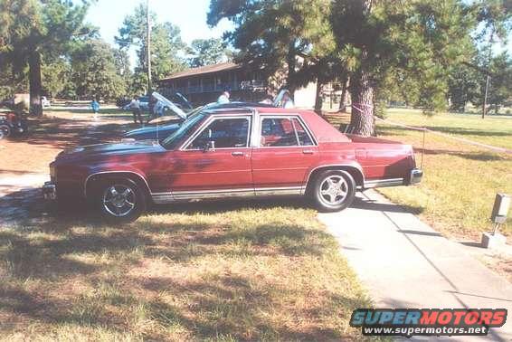 Red sedan with chrome trim parked on a sidewalk, another car with hood up in background