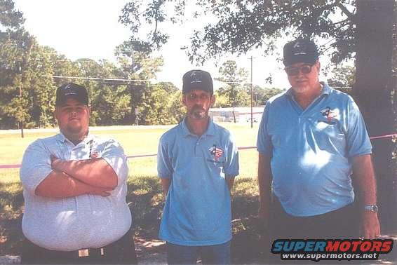 Three men stand outside, two wearing caps. The man on the left has arms crossed
