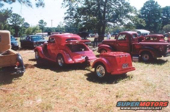 Red coupe with a matching trailer, parked near several trucks outdoors