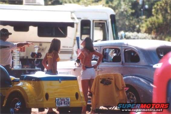 Two women by a vintage car and a yellow trailer with a license plate that reads 86N-MAN. An RV is in the background