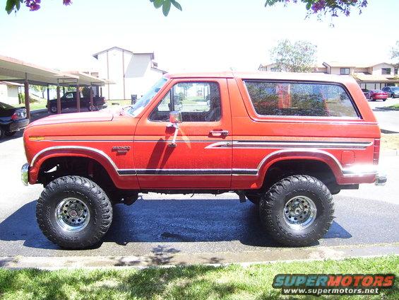 Side view of a red lifted 1986 Ford Bronco with chrome wheels and off-road tires parked outside