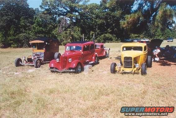 Three hot rod cars parked on grass, one red, one yellow, one brown and black. Trees in background