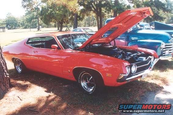 Red 1970s muscle car with hood open, showing engine. Another blue truck is behind it