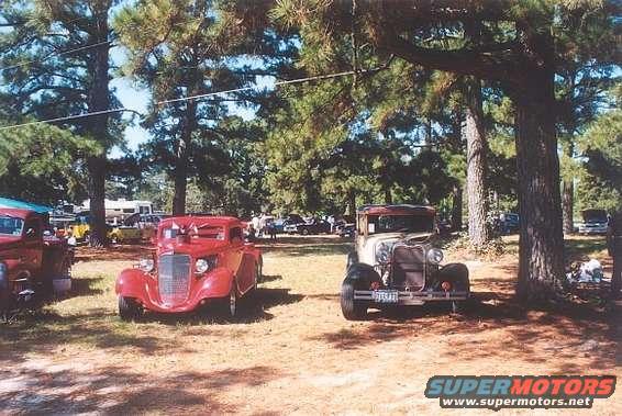 Two old cars parked outside under trees. One is red, the other is tan