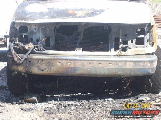 Front bumper and grille area of a vehicle heavily damaged by fire, with exposed radiator and wiring