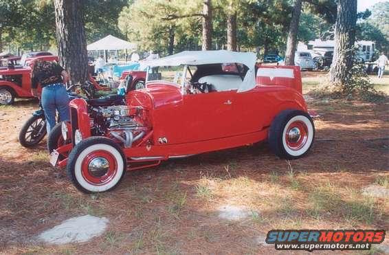 Red hot rod roadster with white convertible top, exposed engine, and whitewall tires. Another red car and a person are visible behind it
