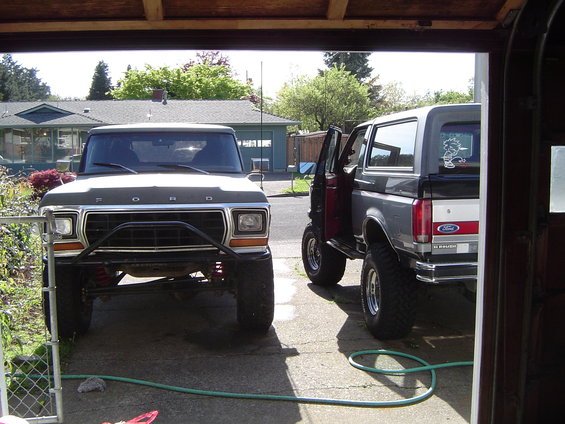 Two Ford Broncos parked in a driveway, one dark gray and one dark brown. The dark gray Bronco has its door open