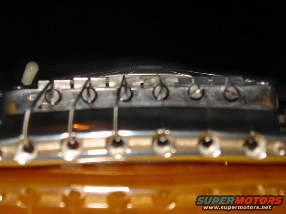 Close-up of an electric guitar bridge with six strings and tuning pegs. Watermark reads SUPERMOTORS