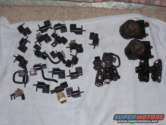 Pile of various black plastic and metal automotive vacuum solenoids and actuators laid out on a white cloth