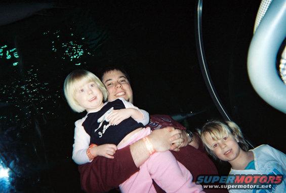 Dad holds a young girl while another girl stands nearby. They are in front of a dark background with glowing green specks, possibly at an aquarium