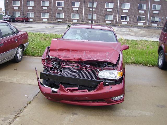 Front view of a red sedan with its hood crumpled and grille damaged, parked next to another red car. A red van is partially visible on the right