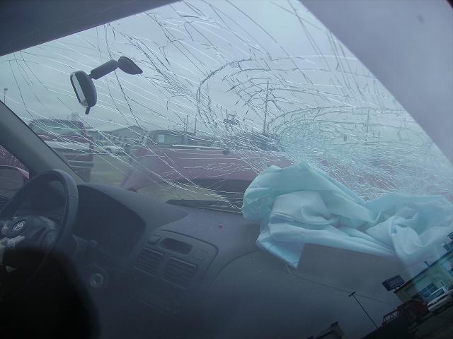 Interior view of a car with a shattered windshield and a deployed airbag. Two other vehicles are visible outside