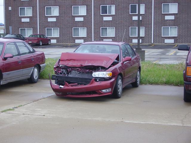 Front end of a red sedan with heavy damage, parked next to another car. Building in background