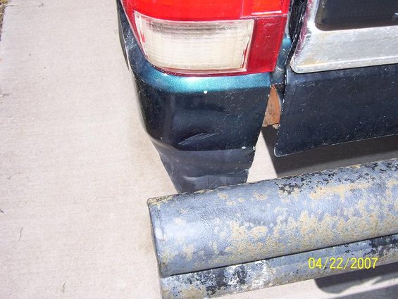 Rear bumper and taillight of a dark green truck, with a rusty metal bar in the foreground showing a date stamp