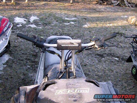 Handlebars and dashboard of a snowmobile, with another snowmobile to the left