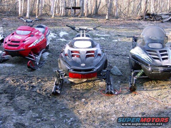 Three snowmobiles parked in a dirt and grass area with trees in the background. The snowmobiles are red, black, and black with green accents