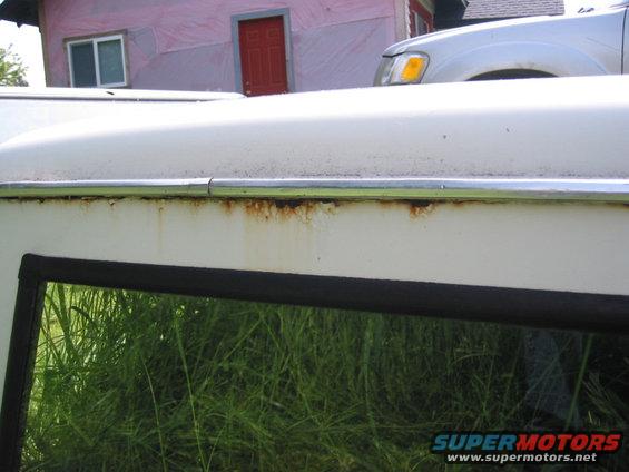Close-up of rusted area on a white vehicle's roofline above a window reflecting grass