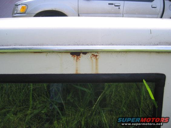 Close-up of rust streaks on a white vehicle's side panel, with another silver vehicle visible above. Grass is reflected in the window