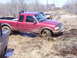 1996 Ford Bronco - A red pickup truck is stuck in deep mud on an off-road trail, with other vehicles visible in the background.
