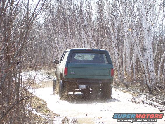 Green 1996 Ford Bronco driving through a muddy trail with trees on either side