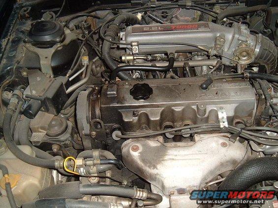 Engine bay of a 1989 Mazda MX-6, showing the 2.2L turbo engine with visible hoses, wiring, and intake manifold