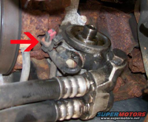 Engine oil filter housing with red arrow pointing to electrical connector. Rusty metal and hoses visible