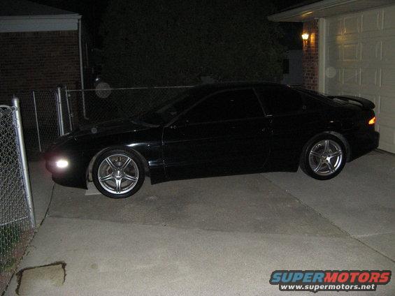 Black coupe parked in driveway at night, side view. Aftermarket wheels and spoiler visible