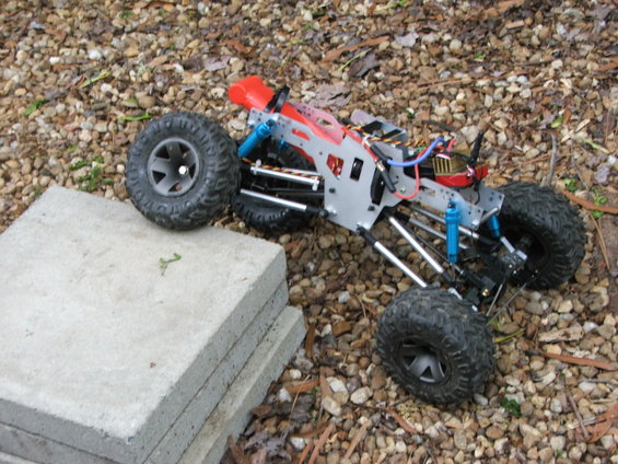 RC rock crawler chassis with large tires, blue shocks, and red bodywork, resting on gravel and concrete slabs