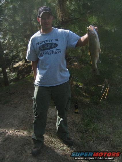 Man in LAFD t-shirt holds a large fish outdoors, with trees and grass in the background