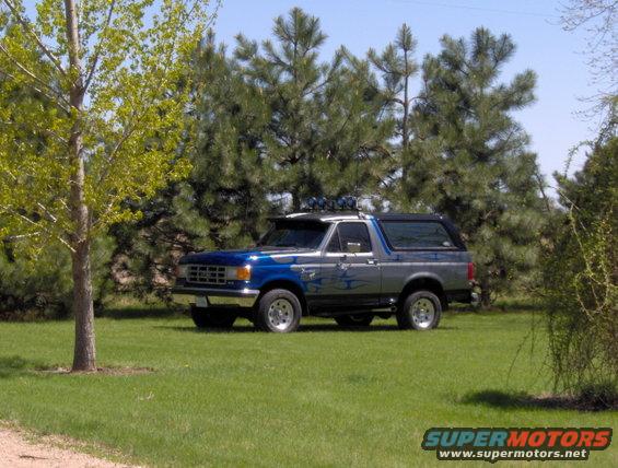 Blue and silver Ford Bronco parked on grass with trees behind it