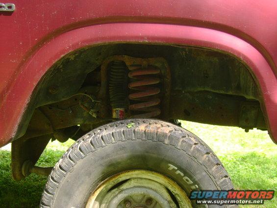 Red Ford Bronco front wheel well with visible suspension components and tire. The body style suggests an 80s model