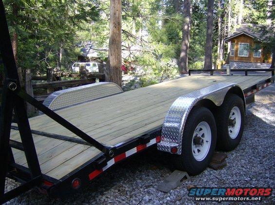 Car trailer with wooden deck and diamond plate fenders, parked on gravel near trees and a cabin