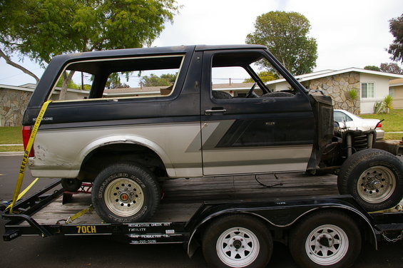 Ford Bronco on a trailer, missing hood and some interior parts. It has a two-tone paint job with a silver lower half and black upper half