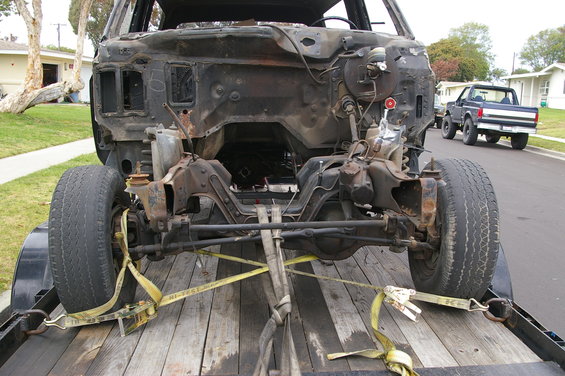 Front clip of a heavily damaged vehicle on a trailer, with a blue pickup truck in the background