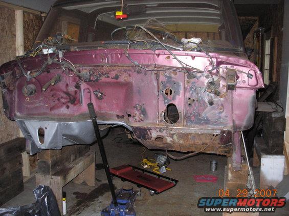 Vehicle firewall disassembled, showing wiring and rust. Supported by wooden blocks and a jack