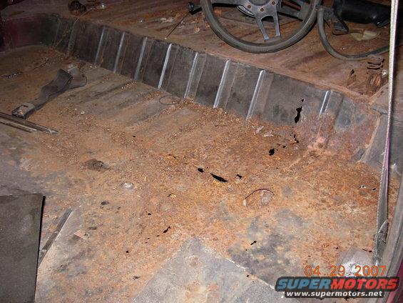 Interior of a rusty vehicle with holes in the floor and a steering wheel visible