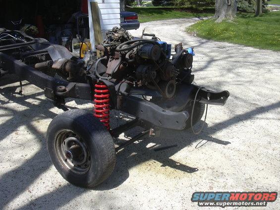 Vehicle chassis with engine and red coil springs, front tire visible, parked on gravel driveway