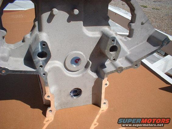 Close-up of an engine block with two mounting holes visible. A circular sticker with text is on the surface