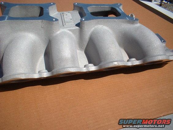 Aluminum intake manifold with four runners and two rectangular openings, resting on a brown surface